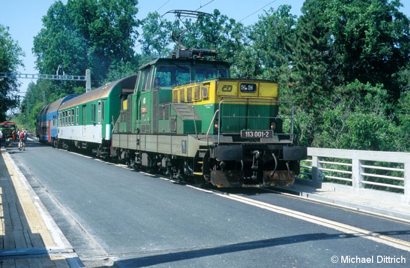 Bild: Eine Eisenbahn auf der Straße? Ja, das gibt es wirklich. 
Die 113 001 fährt gerade aus Bechyne über die Brücke. Neben ihr ist eine Straße wo gerade Bauarbeiten stattfinden.